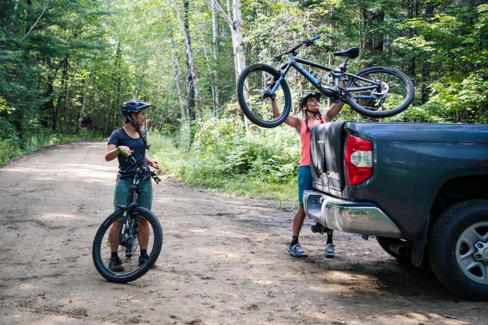 A woman stands astride a mountain bike while a second lifts a bike from the bed of a truck on a dirt road surrounded by trees.