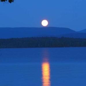 full moon in a blue night sky with mountain sillhuettes on the horizon and the moon reflecting in the lake water