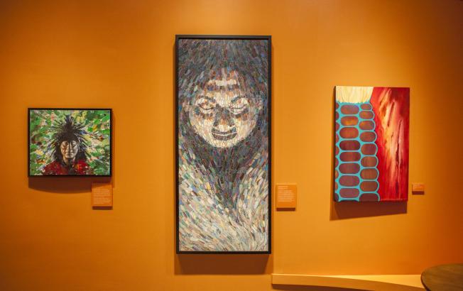 Three paintings by Indigenous artists hang on a deep orange wall in a museum.