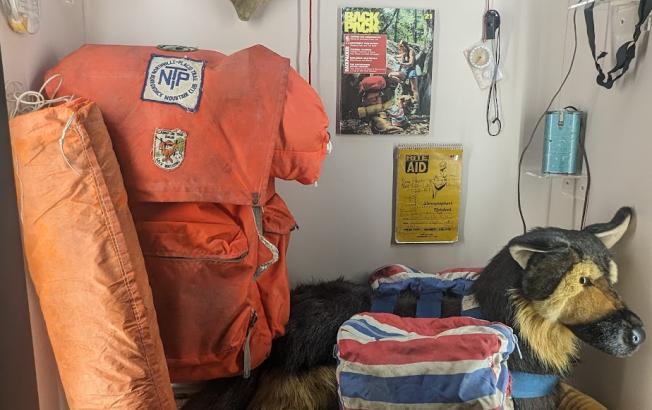 A museum display of historic hiking items, including a well-worn backpack, stuffed dog, and trail guides.