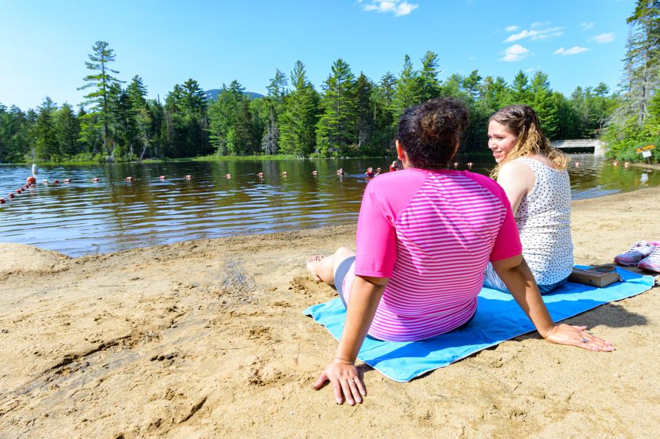Two women in beach clothing sit on a blue towel and look out at a lake on a sunny day.