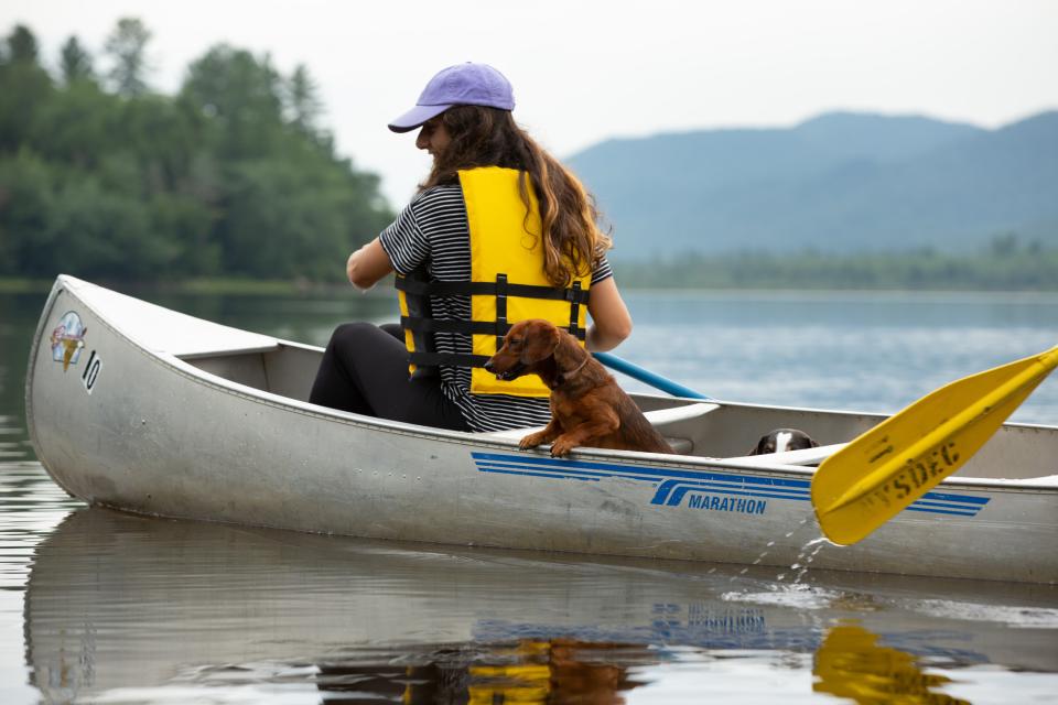 A woman in a hat and yellow lifejacket paddles a canoe with her dachsund dog behind her.