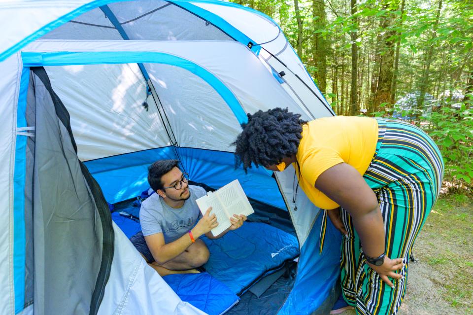 A black woman with yellow shirt and striped pants reads a book held up by a man with glasses in shorts and t shirt in a tent.