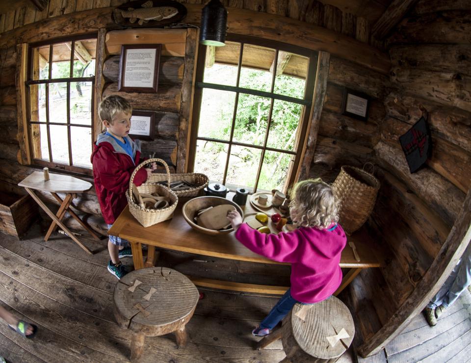 A small log cabin at the Adirondack Experience Museum, with a little boy and little girl playing with the wooden toys and baskets.