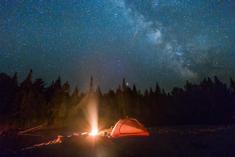 Night time photo of an orange tent glowing from the campfire light nearby. In the background is the black silhouette of pine trees against the night sky filled with stars and with the Milky Way in view.