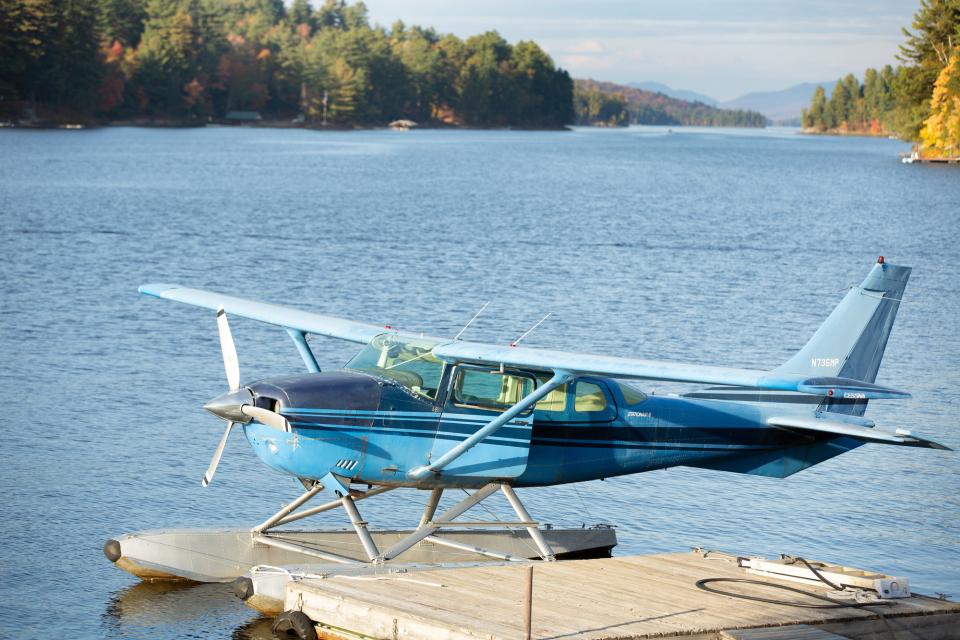 A blue float plane sits on the water in front of a foliage covered backdrop.