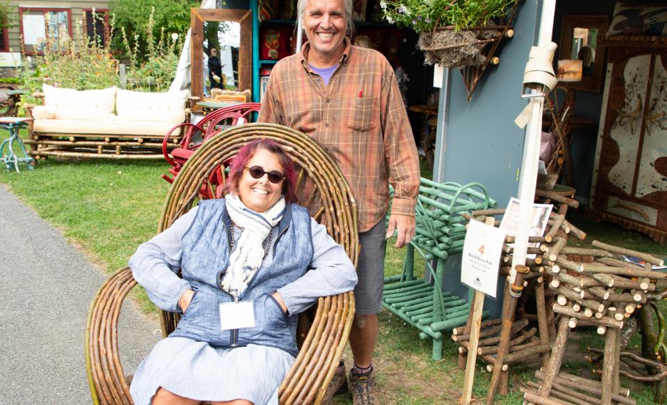 Woman sitting in a rocking chair made of thin branches with a mans standing behind her, both smiling for the photo