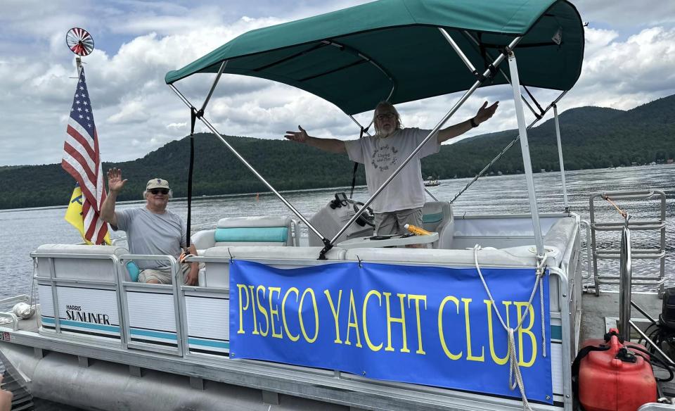 a pontoon boat with two men on board, an American flag flying from a pole, with a banner on the side that reads Piseco Yacht Club