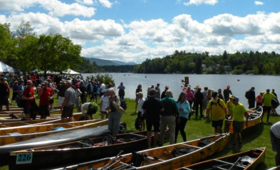 Large group of people wait on the green by the water with their canoes