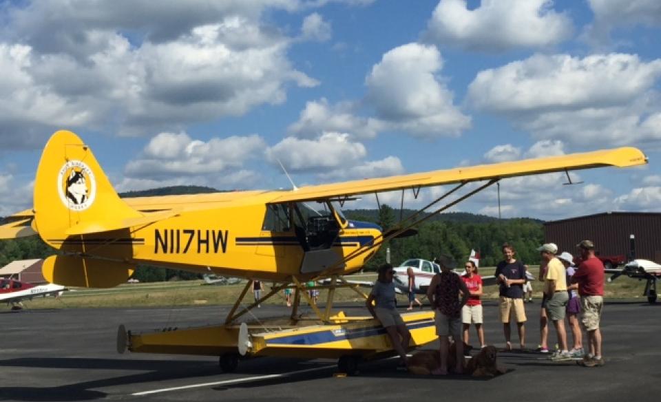 A yellow small plane parked at the airport with several people standing around it looking at it.