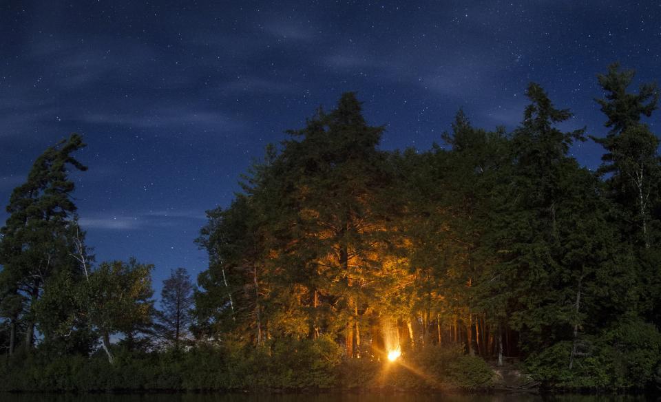 a phot taken at night with a dark blue sky and black silluettes of trees and a small gleam of light of a campfire glowing from the trees