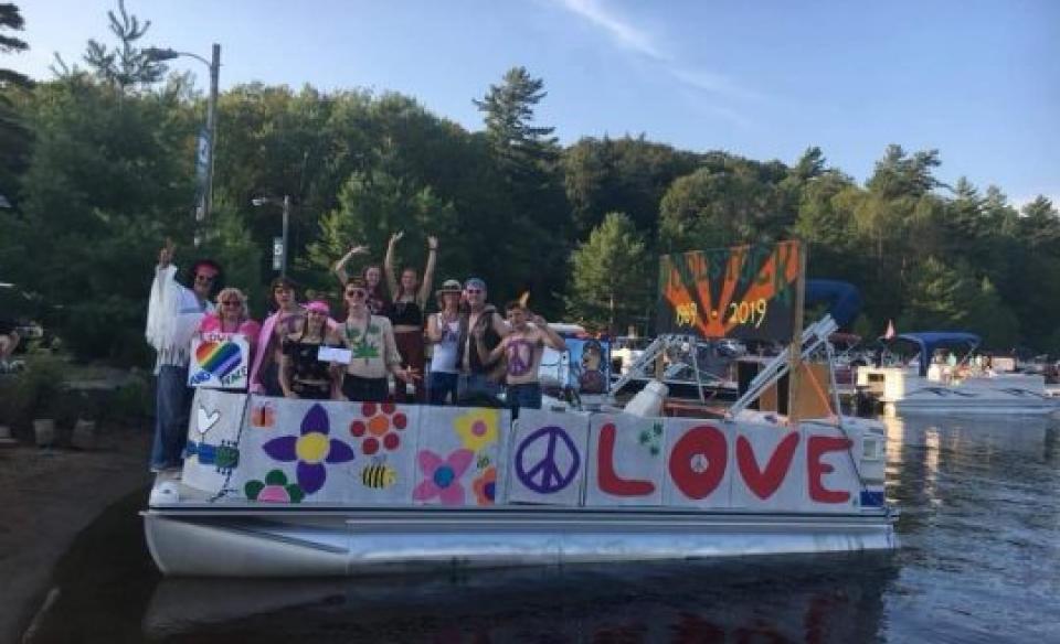 a pontoon boat with many people on board for a boat parade, their boat decorated with a banner of flowers, a peace sign, and the word love